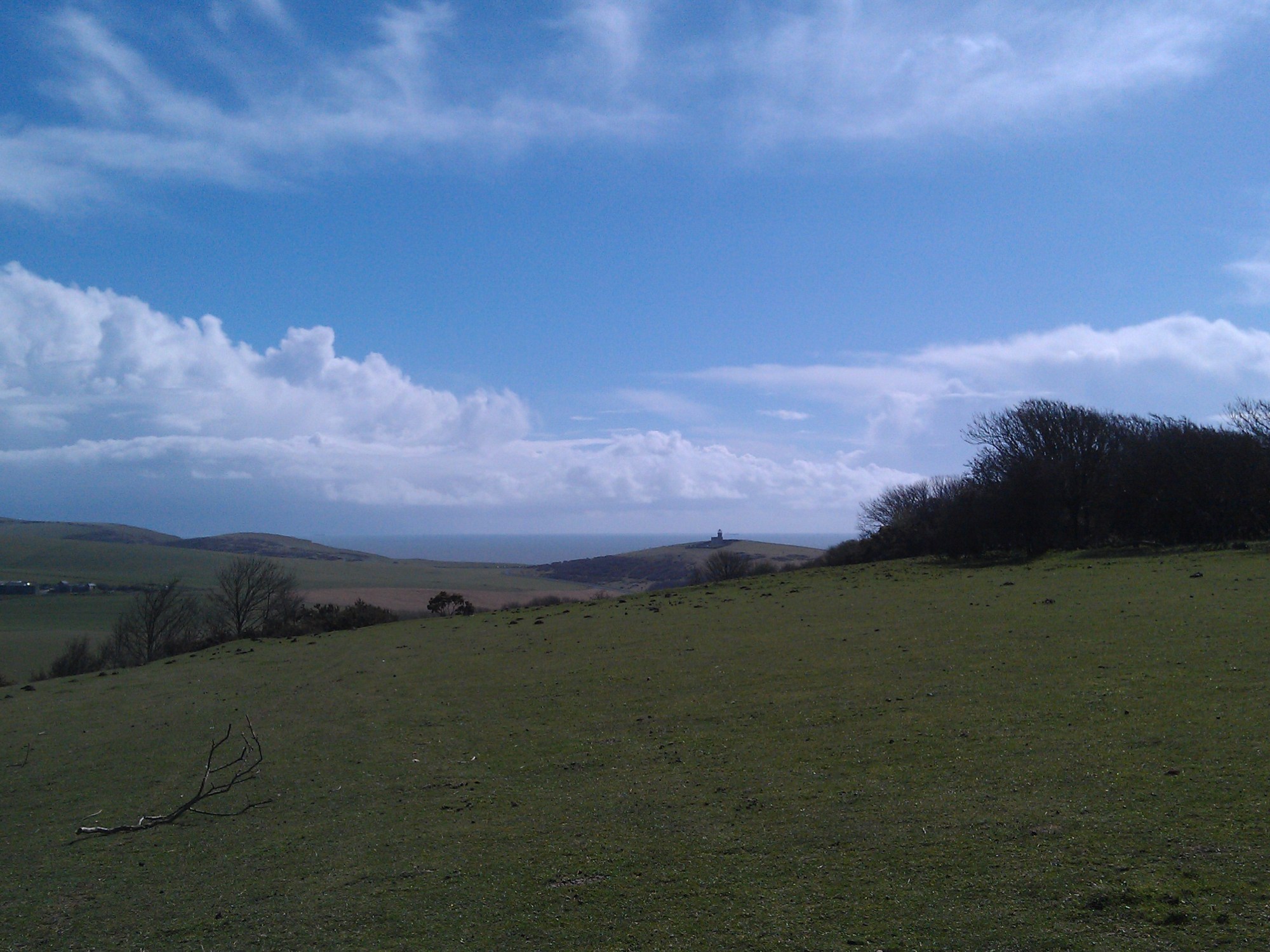 Photo from the South Downs. In the foreground rolling downs below a bright blue sky. In the background you can just make out Belle Toute lighthouse and the sea beyond it.