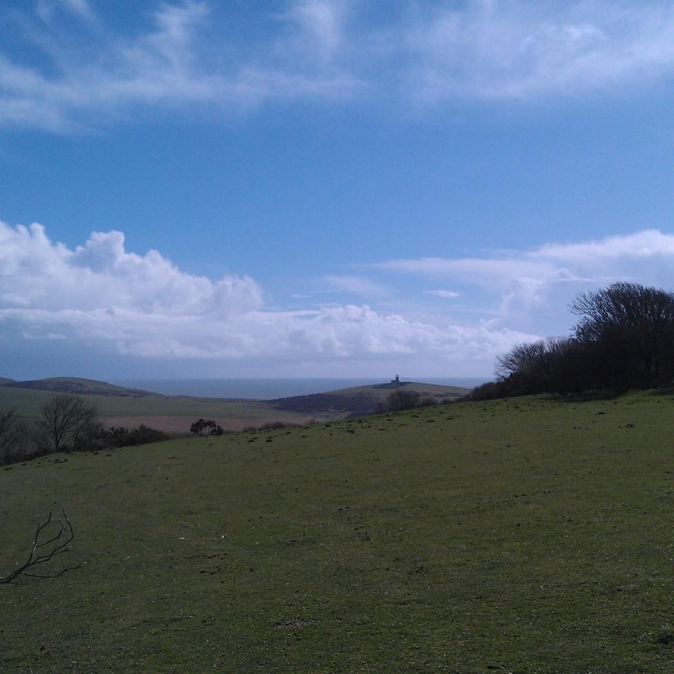 Photo from the South Downs. In the foreground rolling downs below a bright blue sky. In the background you can just make out Belle Toute lighthouse and the sea beyond it.
