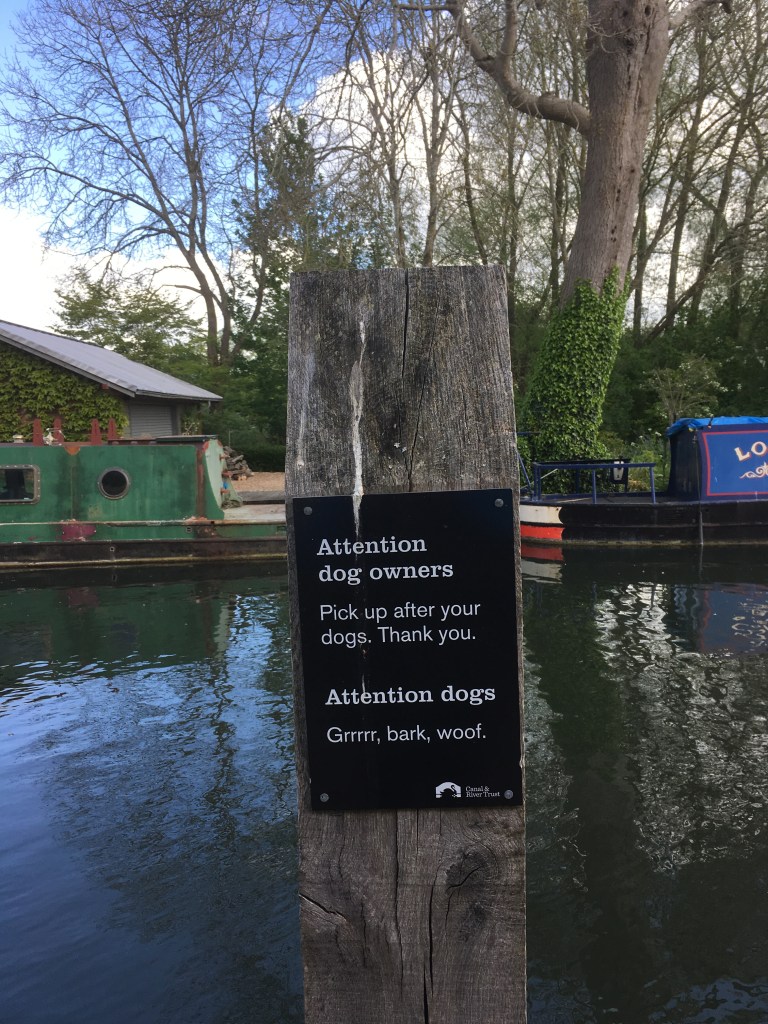 A sign on the Kennet and Avon Canal. it says "Attention dog owners. Pick up after your dogs. Thank you. Attention dogs. Grrrr, bark, woof."