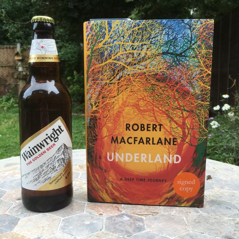 A photo of a bottle of Wainwright Golden Beet and a copy of Underland by Robert Macfarlane on a garden table 