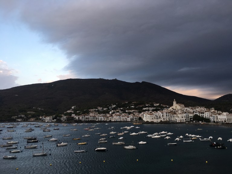 A photo of a harbour.  Small boats dotted across the see and the white buildings of a town on the far hillside.  Above dark clouds disappear over surrounding hills as blue skies move in from the left.